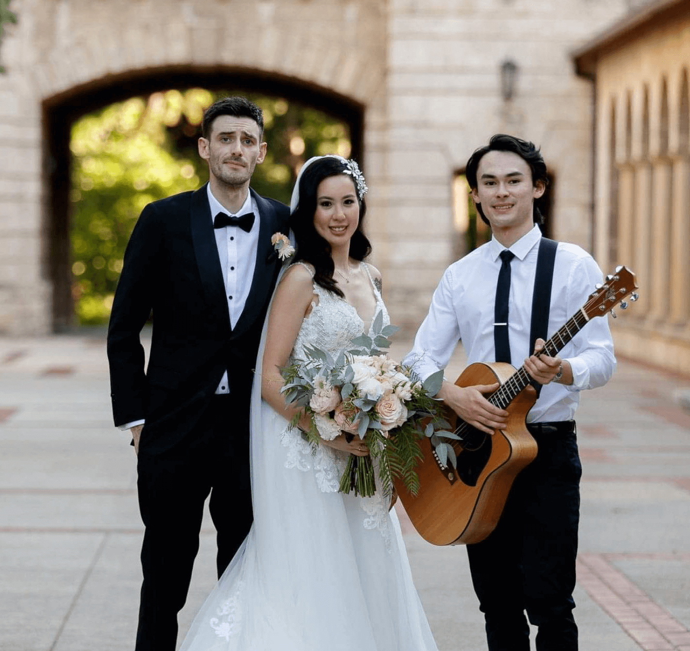 A person in a wedding gown, a person in a suit, and a person holding an acoustic guitar pose outdoors by a stone archway.
