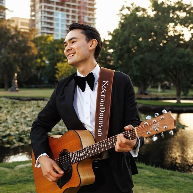 Man in a black tuxedo playing acoustic guitar outdoors near a pond and trees