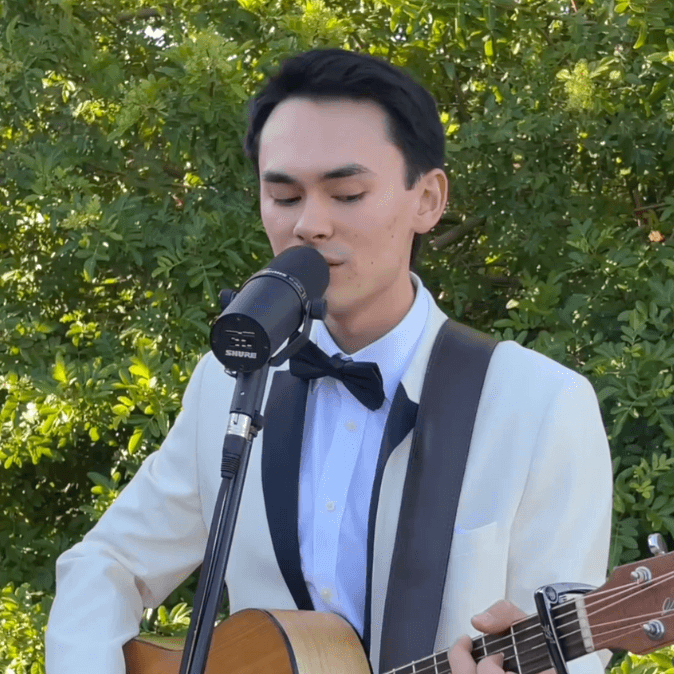 A person in a white tuxedo and black bow tie playing an acoustic guitar and singing into a Shure microphone outdoors.
