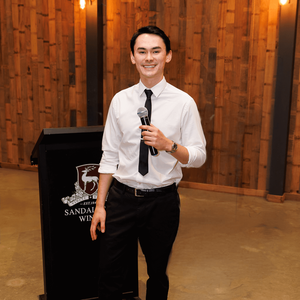 A person in a white dress shirt and black tie holds a microphone at a podium in a room with wood-paneled walls.