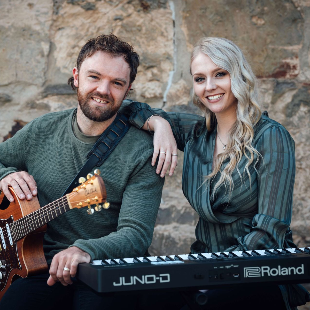 Man with guitar and woman with keyboard, smiling, seated outdoors.