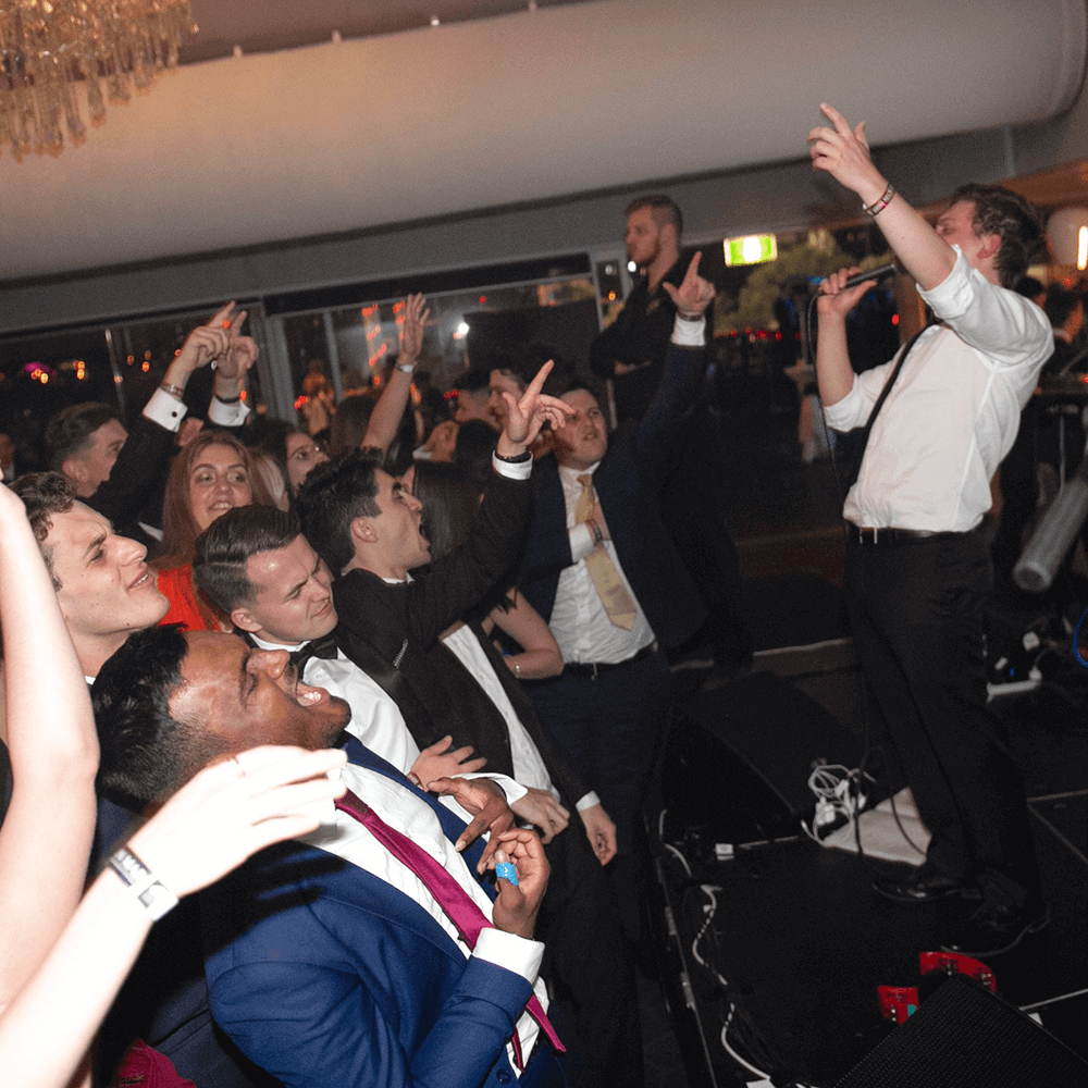 A performer sings into a microphone while a crowd of people in formal attire cheer with arms raised at an indoor event.
