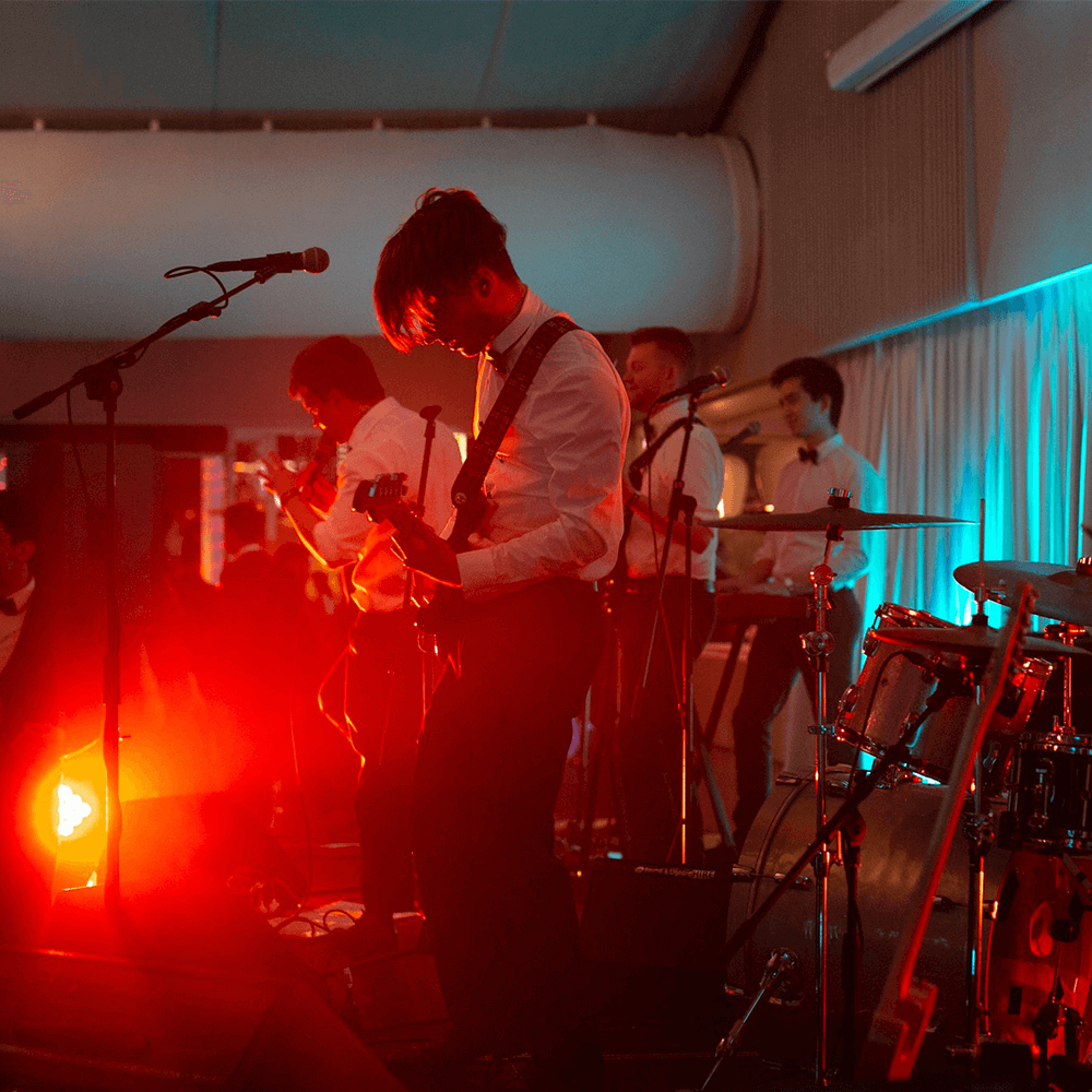 A band in white dress shirts performing on stage under dramatic red lighting, featuring guitars and drums.