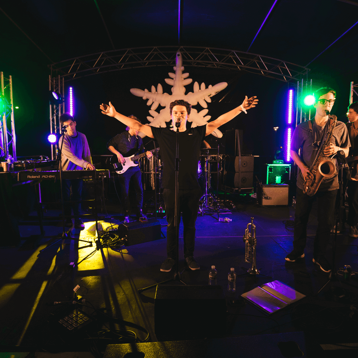A band performs on stage under a large snowflake decoration, with a lead singer center stage, arms spread wide.