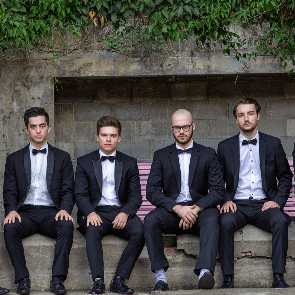 Four men in dark suits and bow ties sit side-by-side on a stone bench against a textured brick wall with greenery.