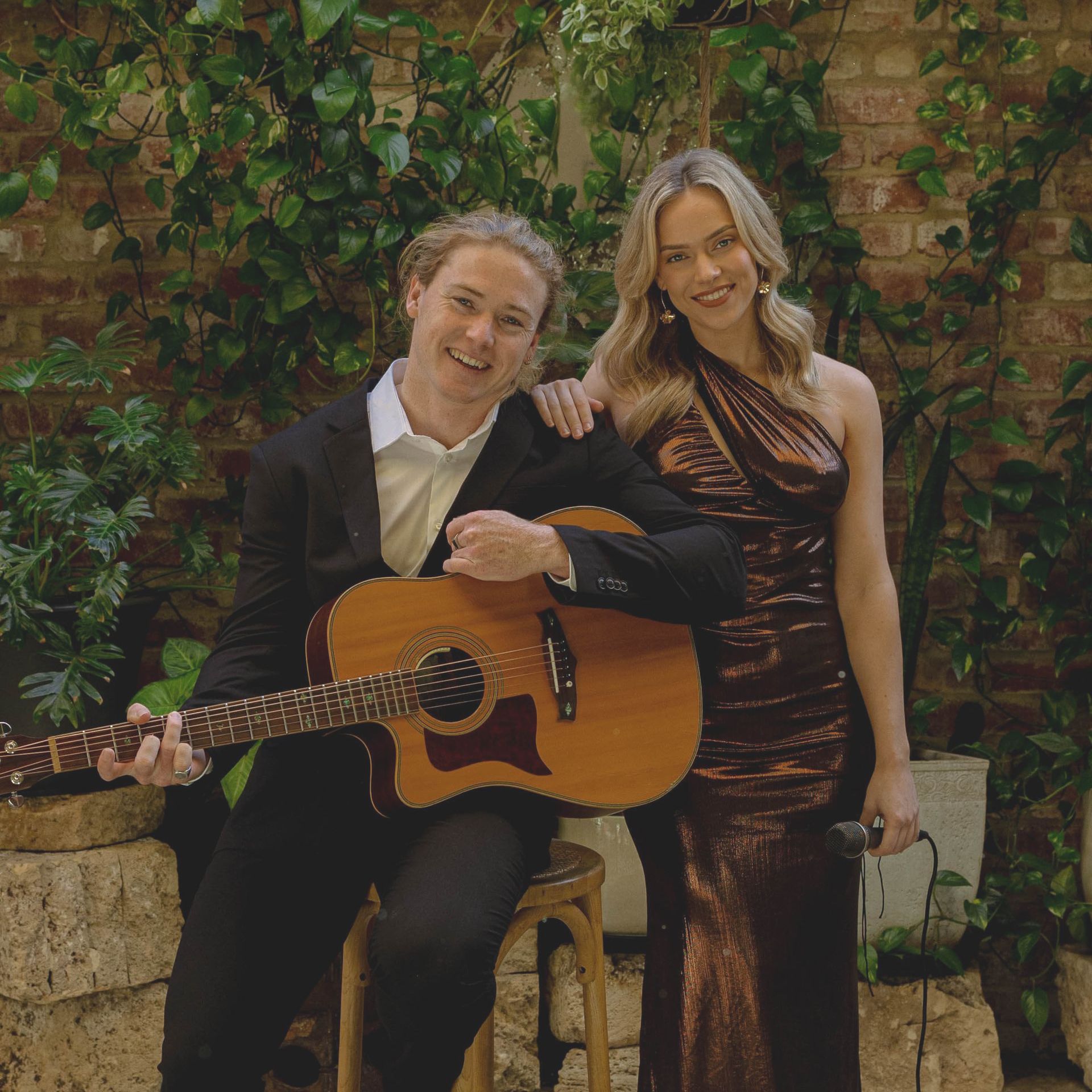 Acoustic duo: man with guitar, woman in bronze dress; brick wall and greenery background.