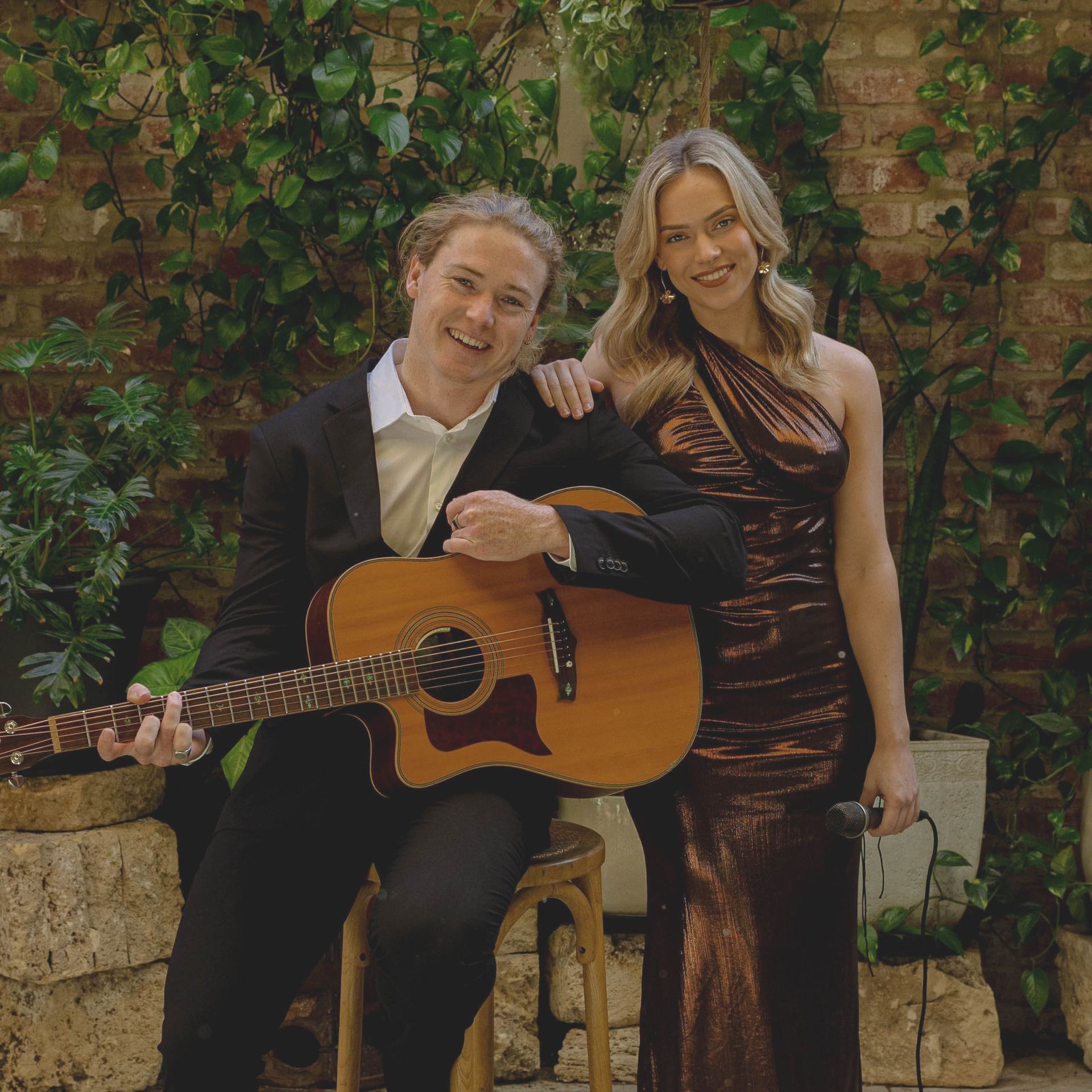 Man with guitar and woman in formal wear pose in front of greenery.