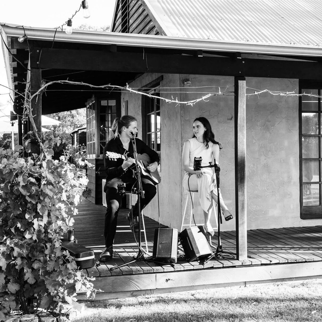 Two musicians performing on a porch, one playing guitar, the other holding a microphone.