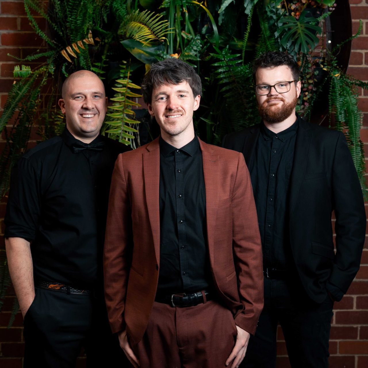 Three men in formal attire posing in front of a brick wall with greenery.
