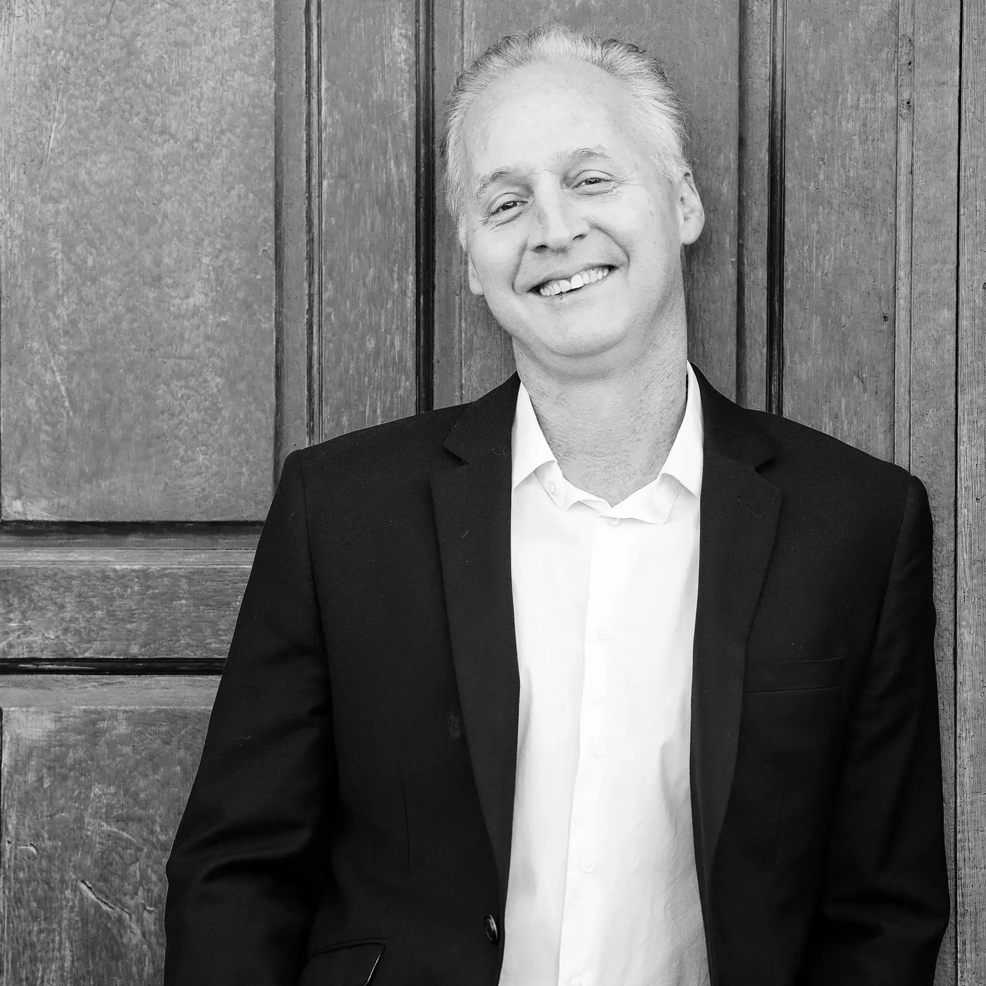 A smiling person in a dark suit jacket and white shirt posing in front of a wooden door.