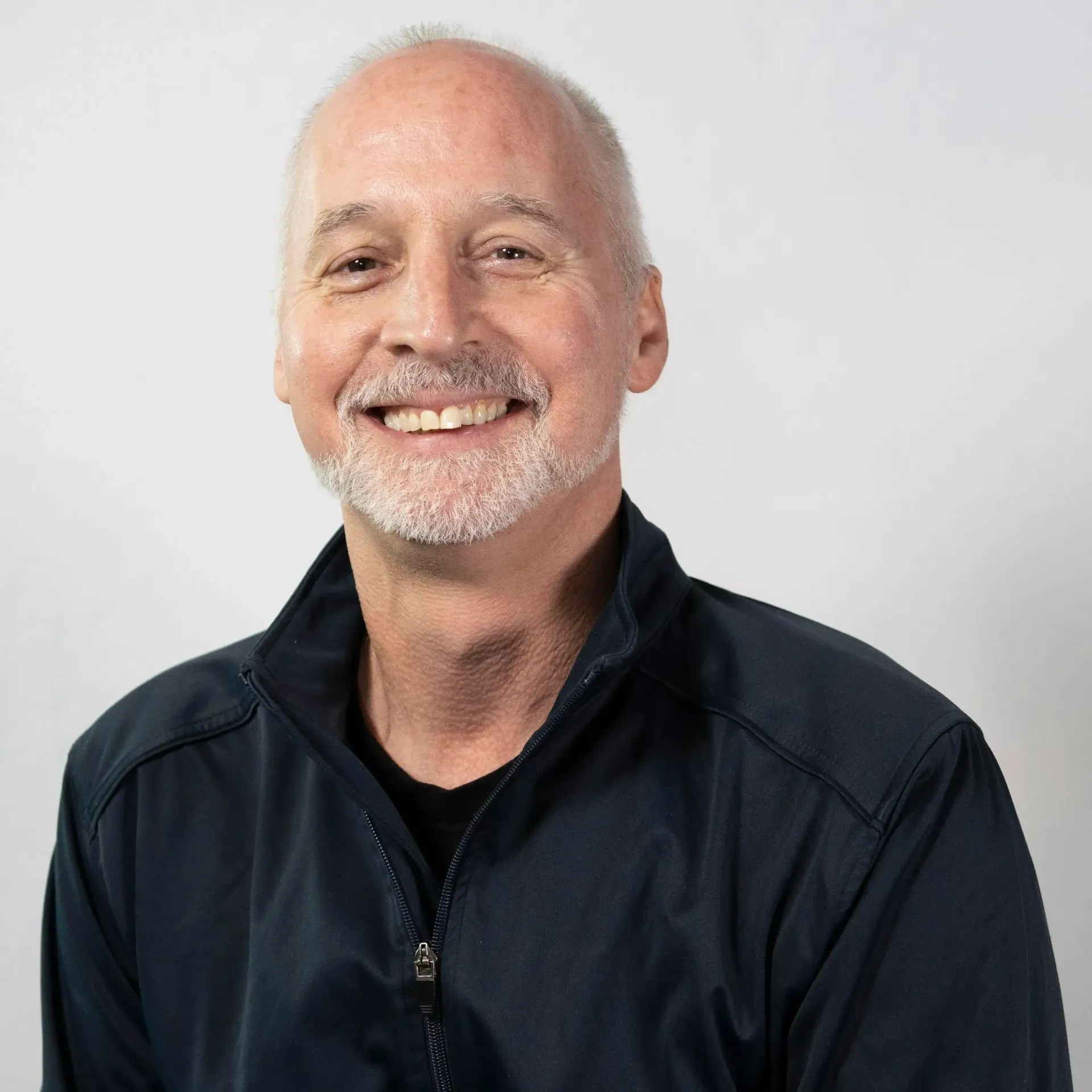 A smiling, middle-aged person with short, light hair and a goatee, wearing a dark blue zip-up jacket against a white wall.