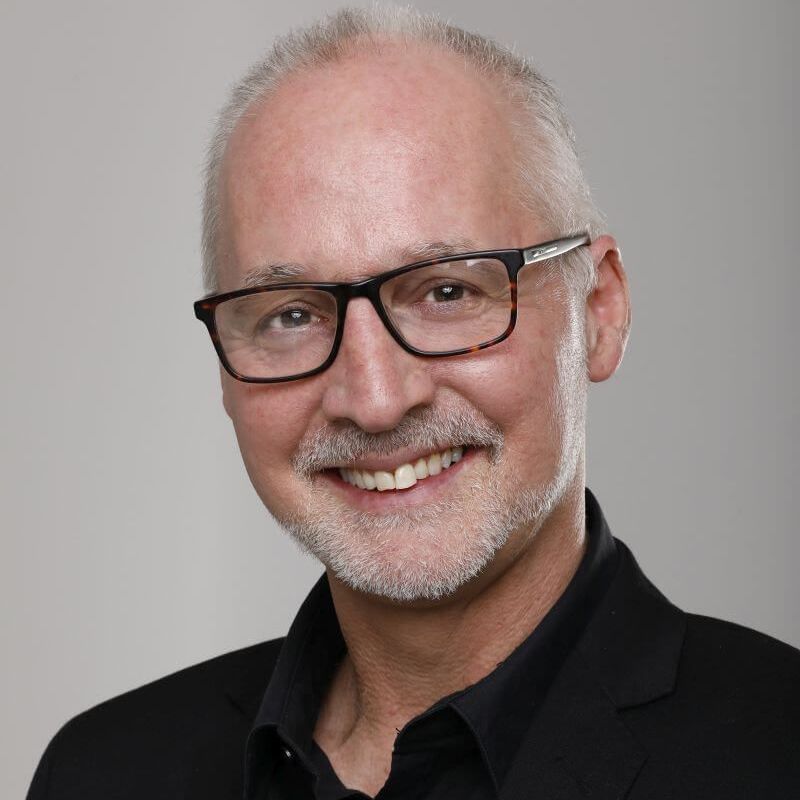 A person with short grey hair and a beard, wearing glasses and a black blazer, smiling against a plain grey background.