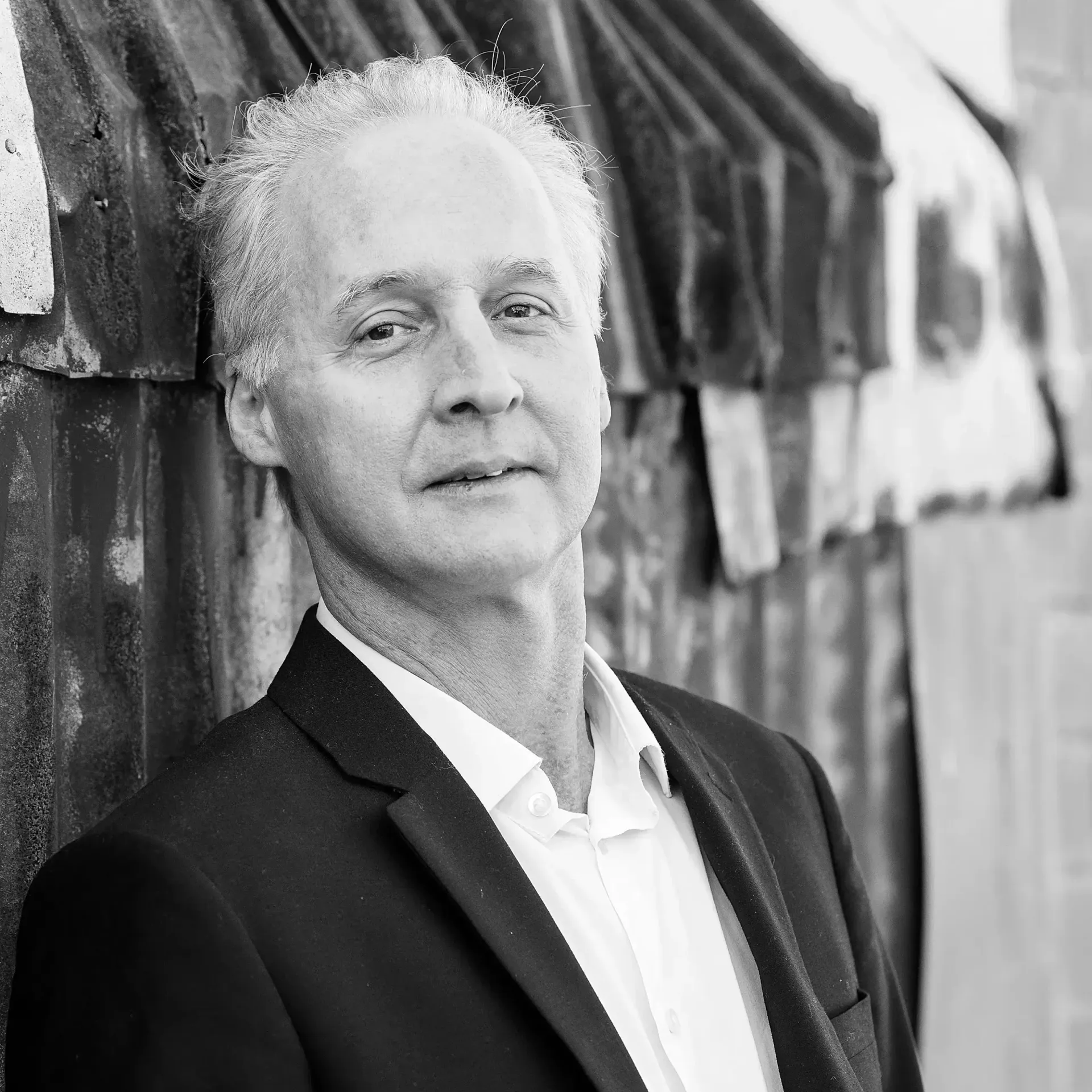 A person in a suit and collared shirt leaning against a textured metal wall in a black and white portrait.