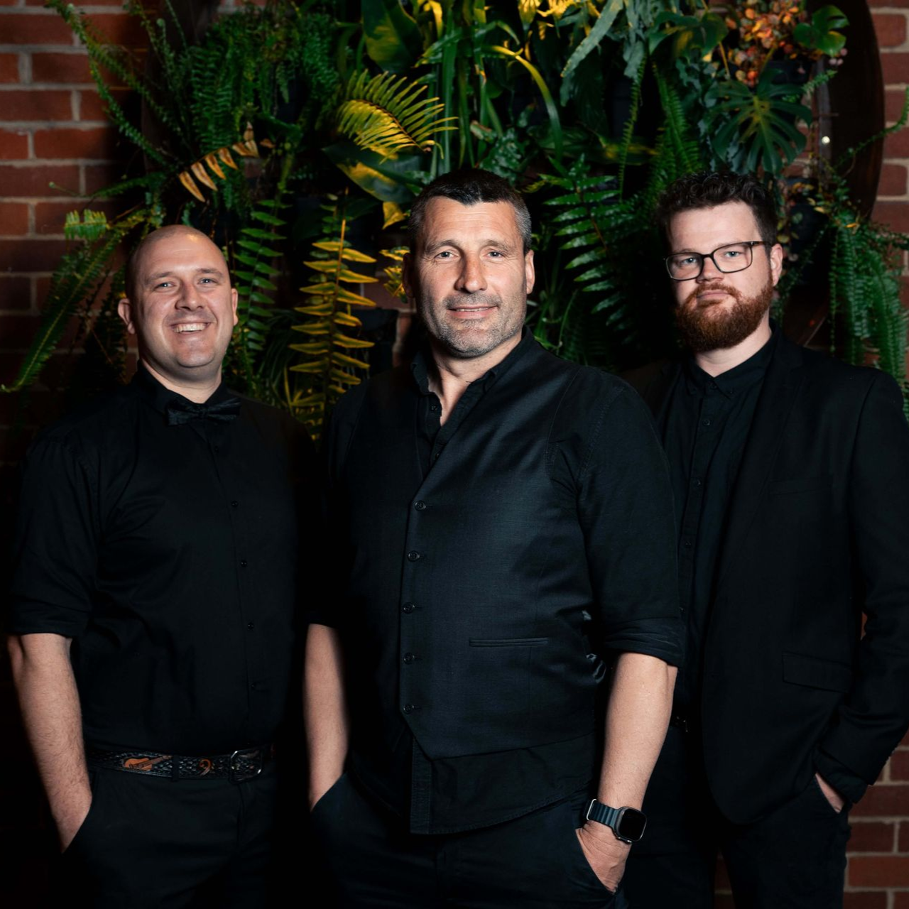 Three men in black outfits posing in front of a brick wall and foliage.