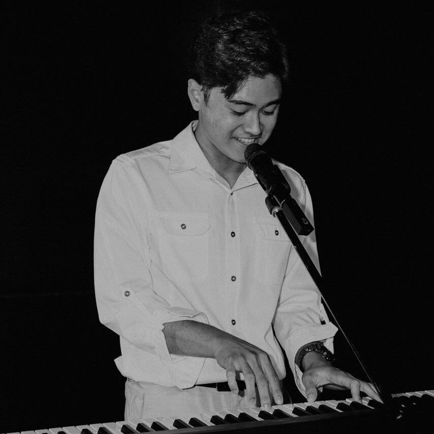Man playing a keyboard and singing into a microphone on stage. Black and white.