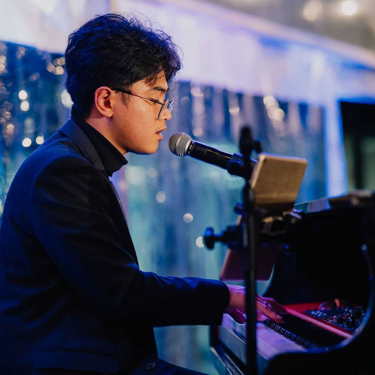 Man playing piano, singing into microphone. Wearing glasses, black suit, and turtleneck. Performing indoors, blue-lit background.