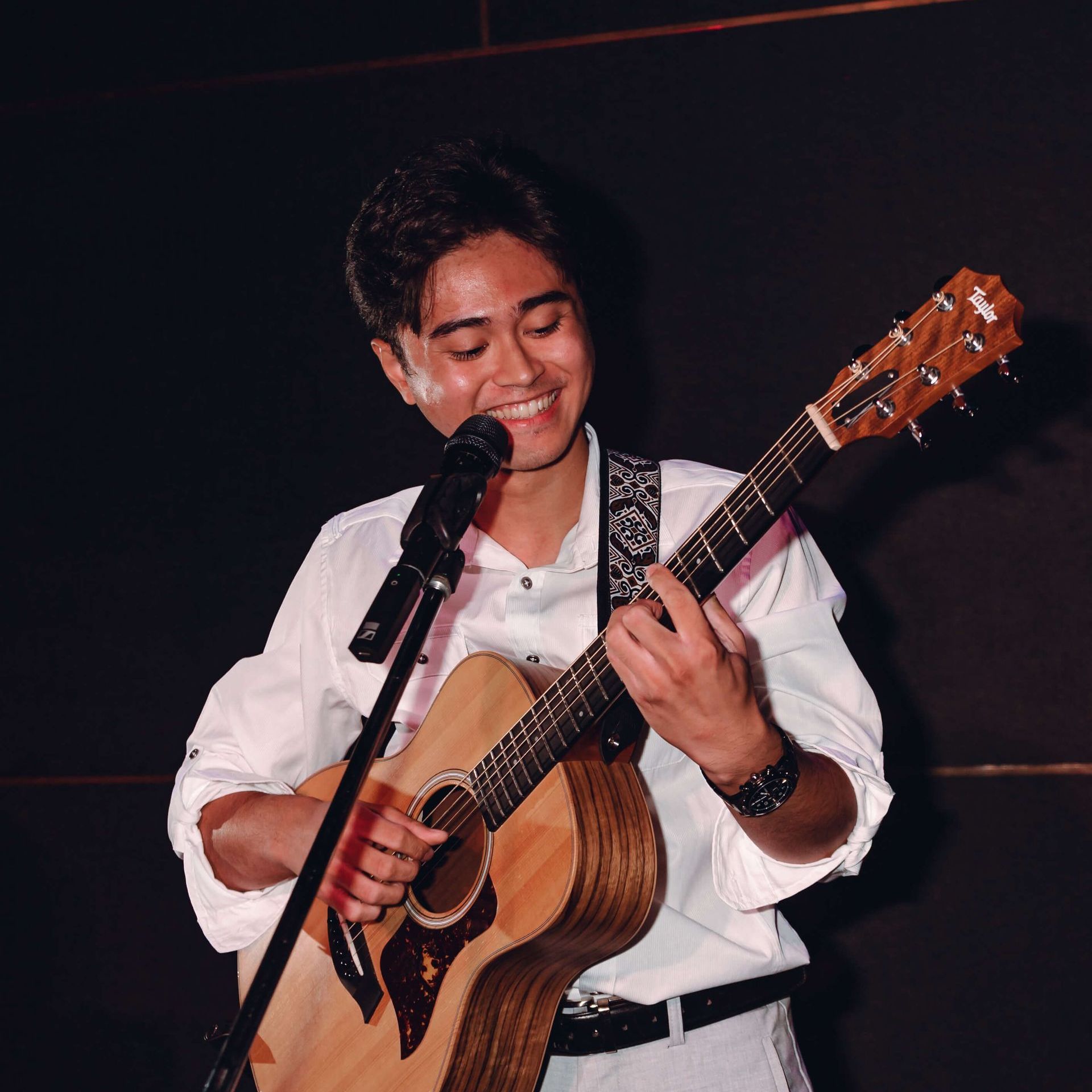 Man smiling, playing acoustic guitar and singing into a microphone on stage, wearing a white shirt.