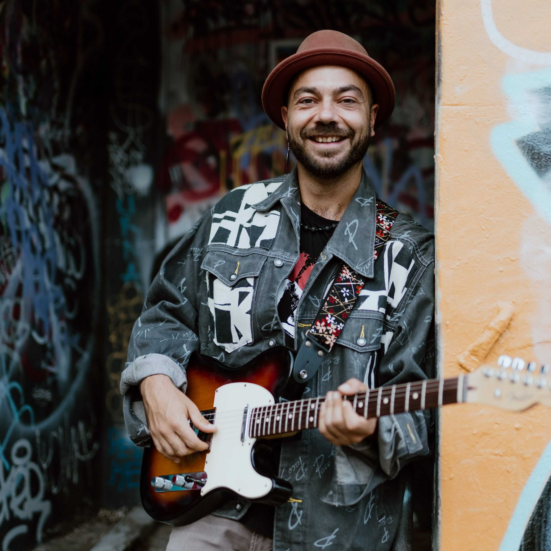 Man holding guitar, smiling, wearing hat and decorated jacket, leaning in graffiti-covered doorway.