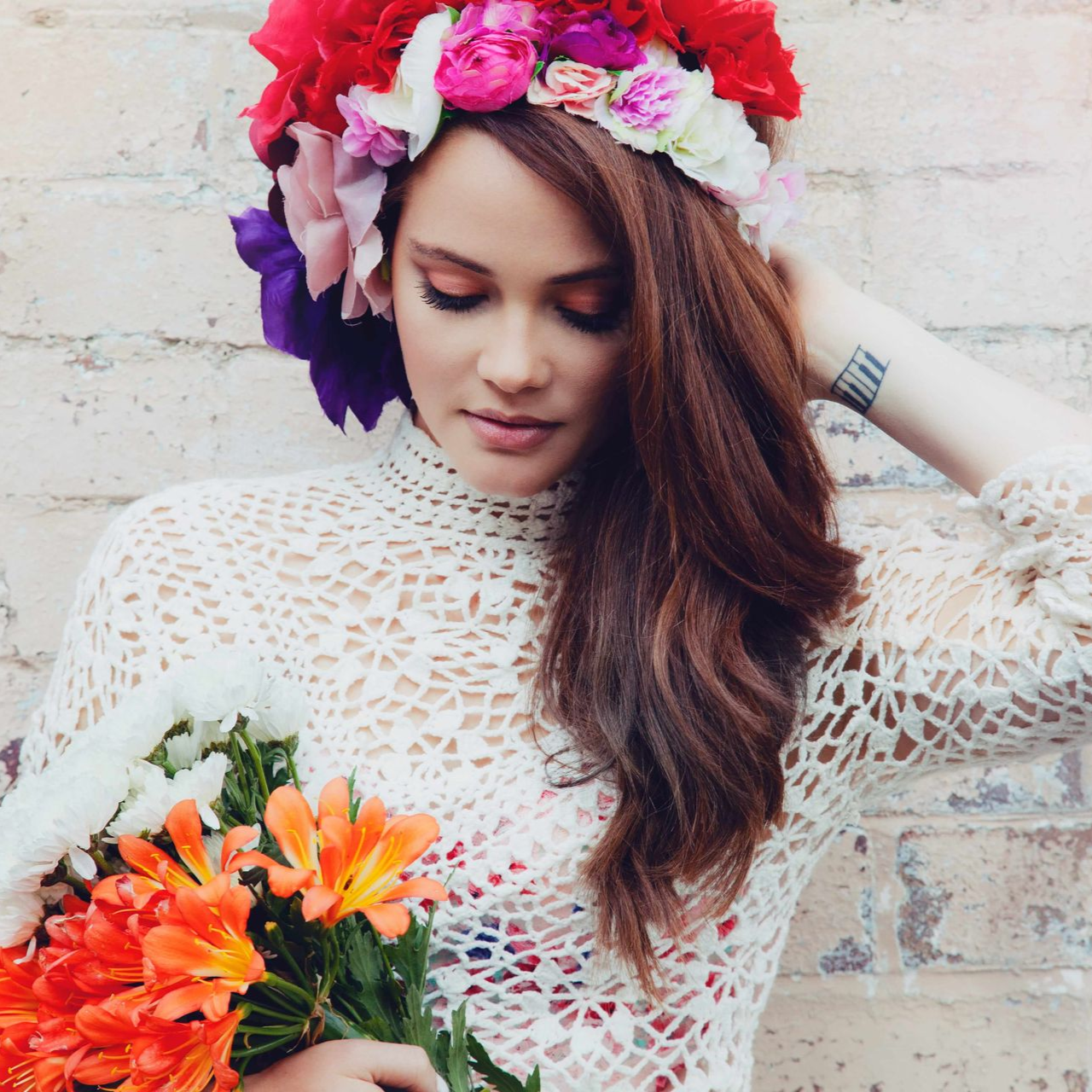 Woman with floral crown and crochet top holding orange flowers, looking down.