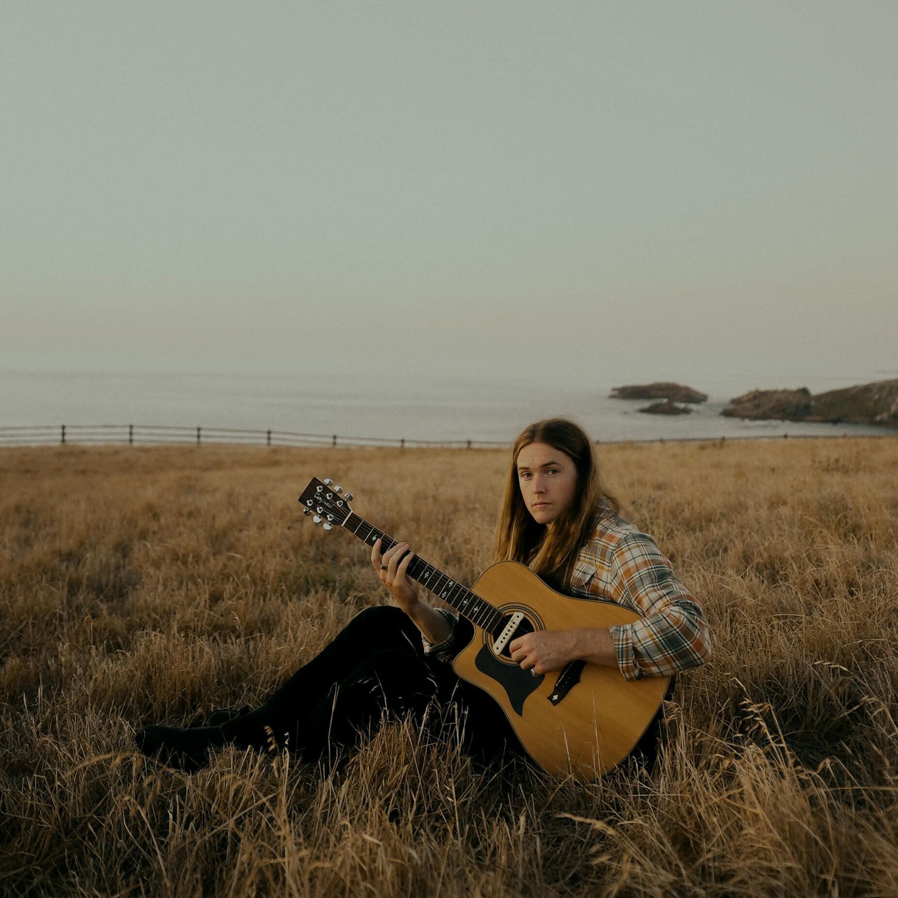 Person sits in tall grass, playing acoustic guitar. Ocean and fog in the background.