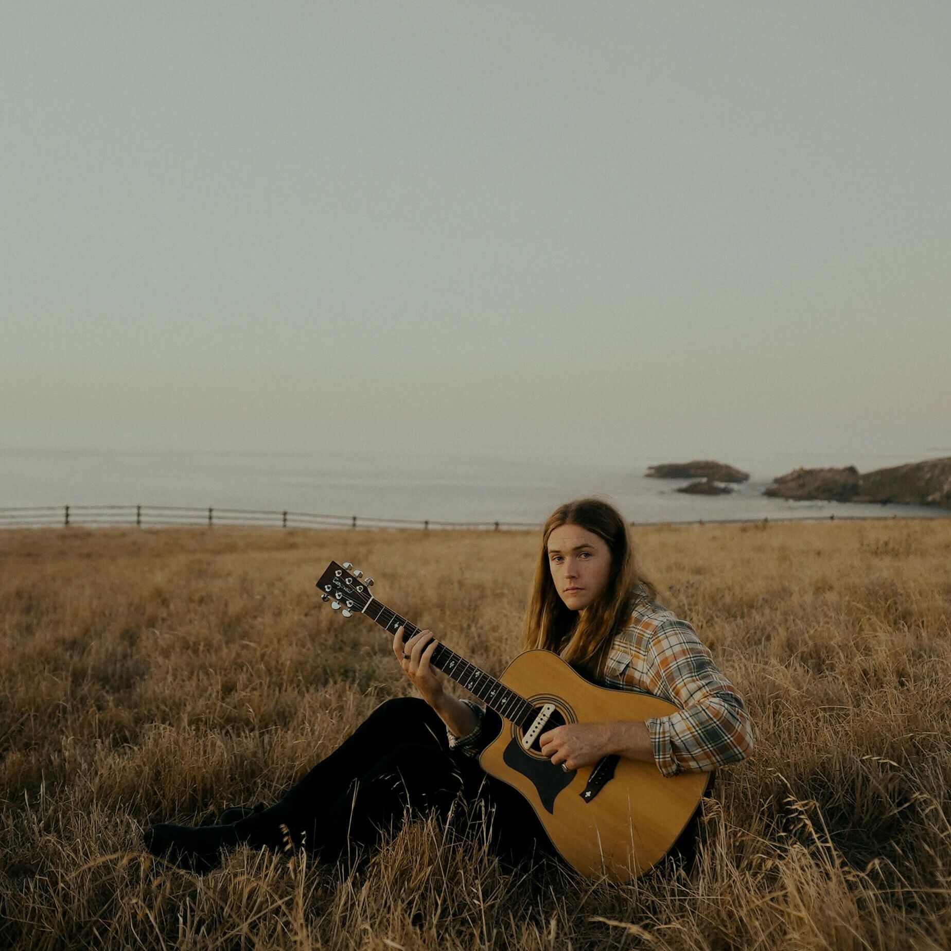 Person playing acoustic guitar in a field, ocean and fence in the background, overcast sky.