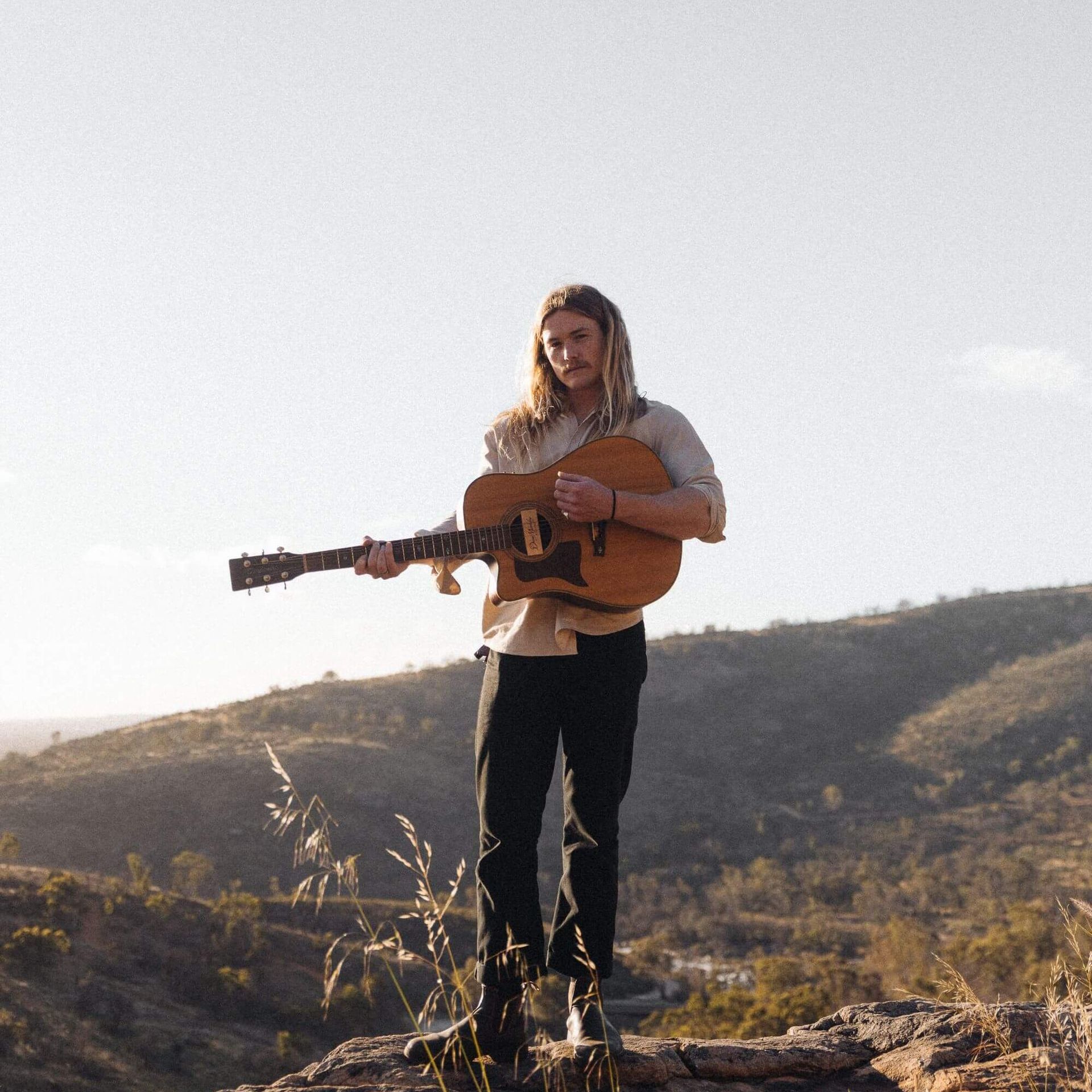 Man with dreadlocks playing guitar on a hilltop, mountains in the background. Sunny day.
