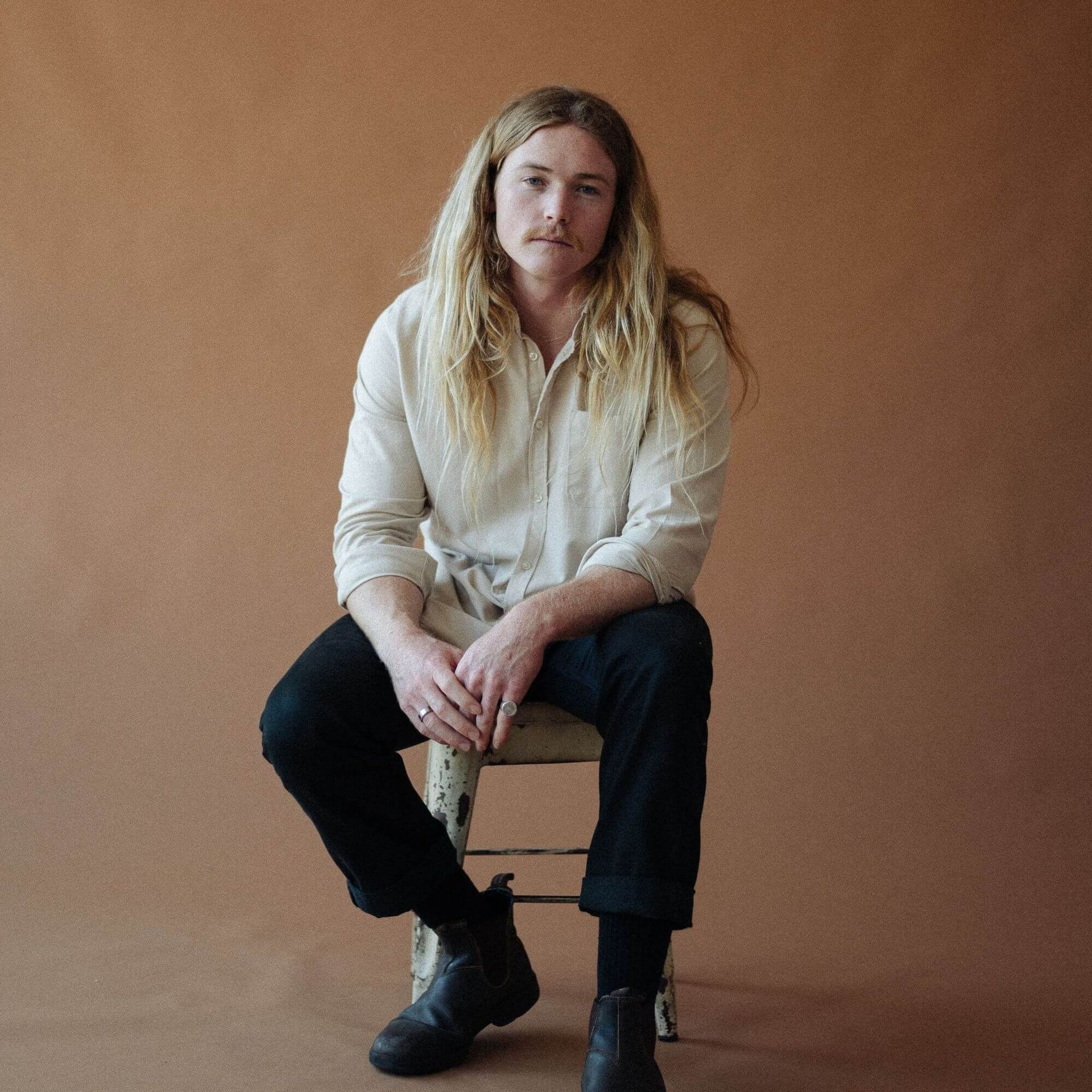 Man with long blonde hair, seated on a chair against a brown backdrop. He wears a button-up shirt, black pants, and boots.
