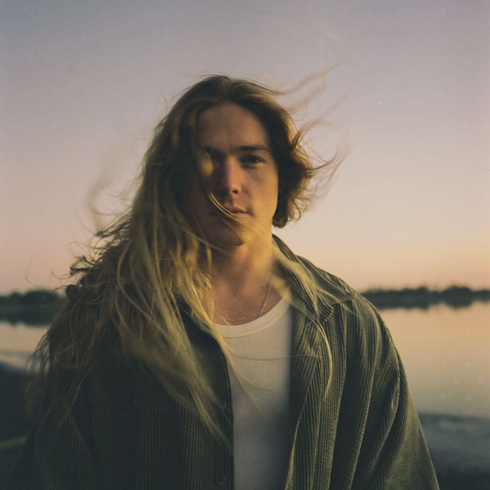 Man with long blonde hair, gazing at the camera outdoors. Wearing white undershirt, green jacket; sunset backdrop.