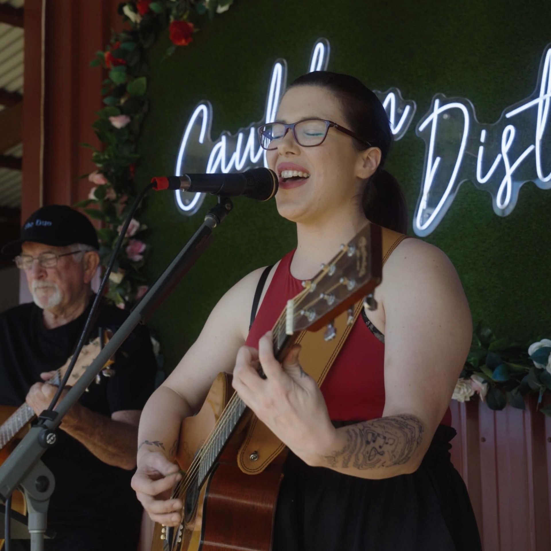 Woman singing and playing guitar on stage, neon sign in background.