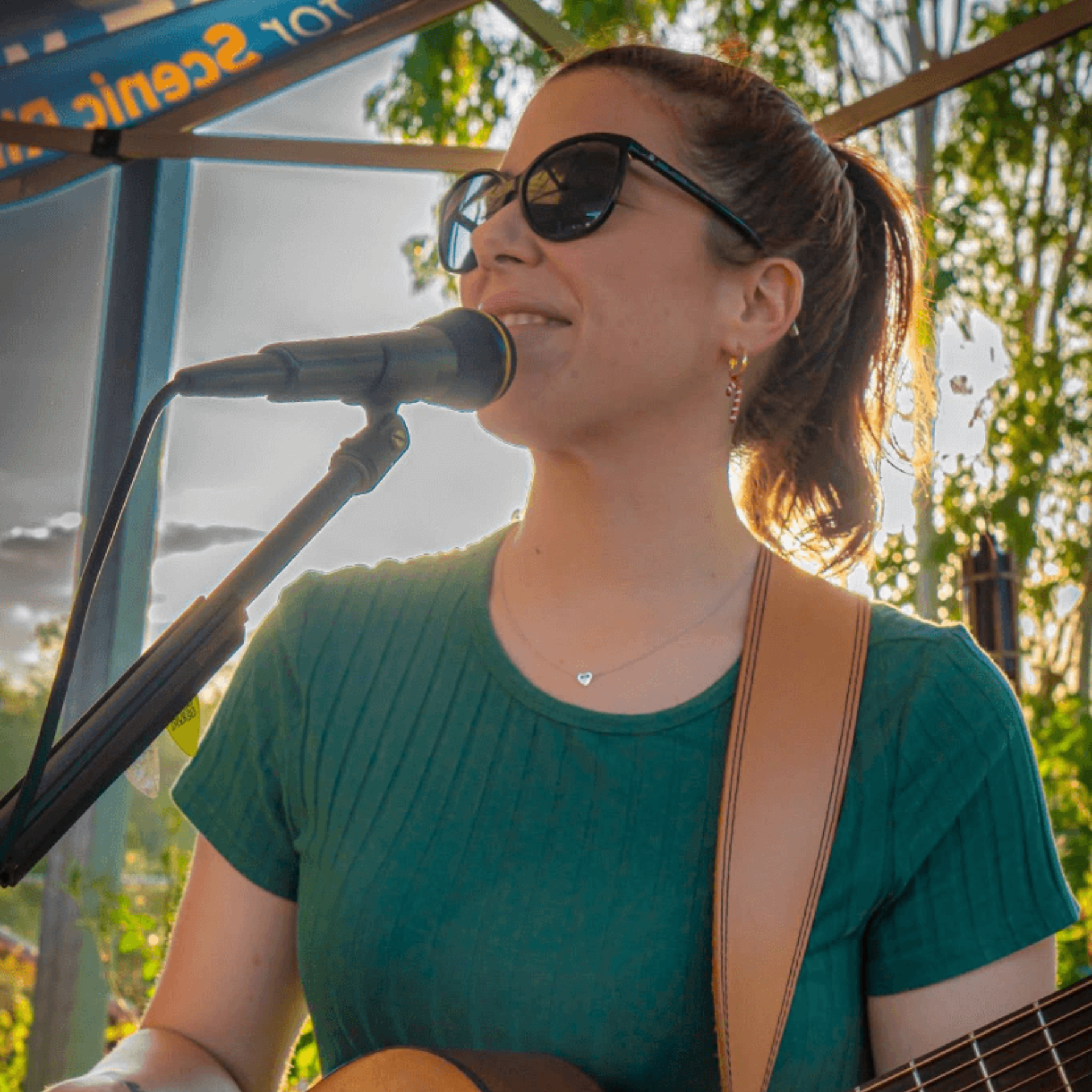 Woman with sunglasses singing and playing guitar outdoors.