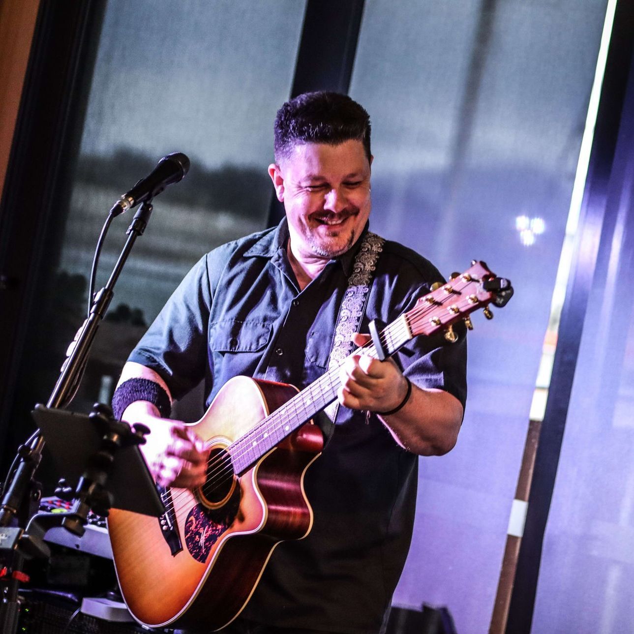 Man playing acoustic guitar, smiling, performing onstage with microphone and tablet.