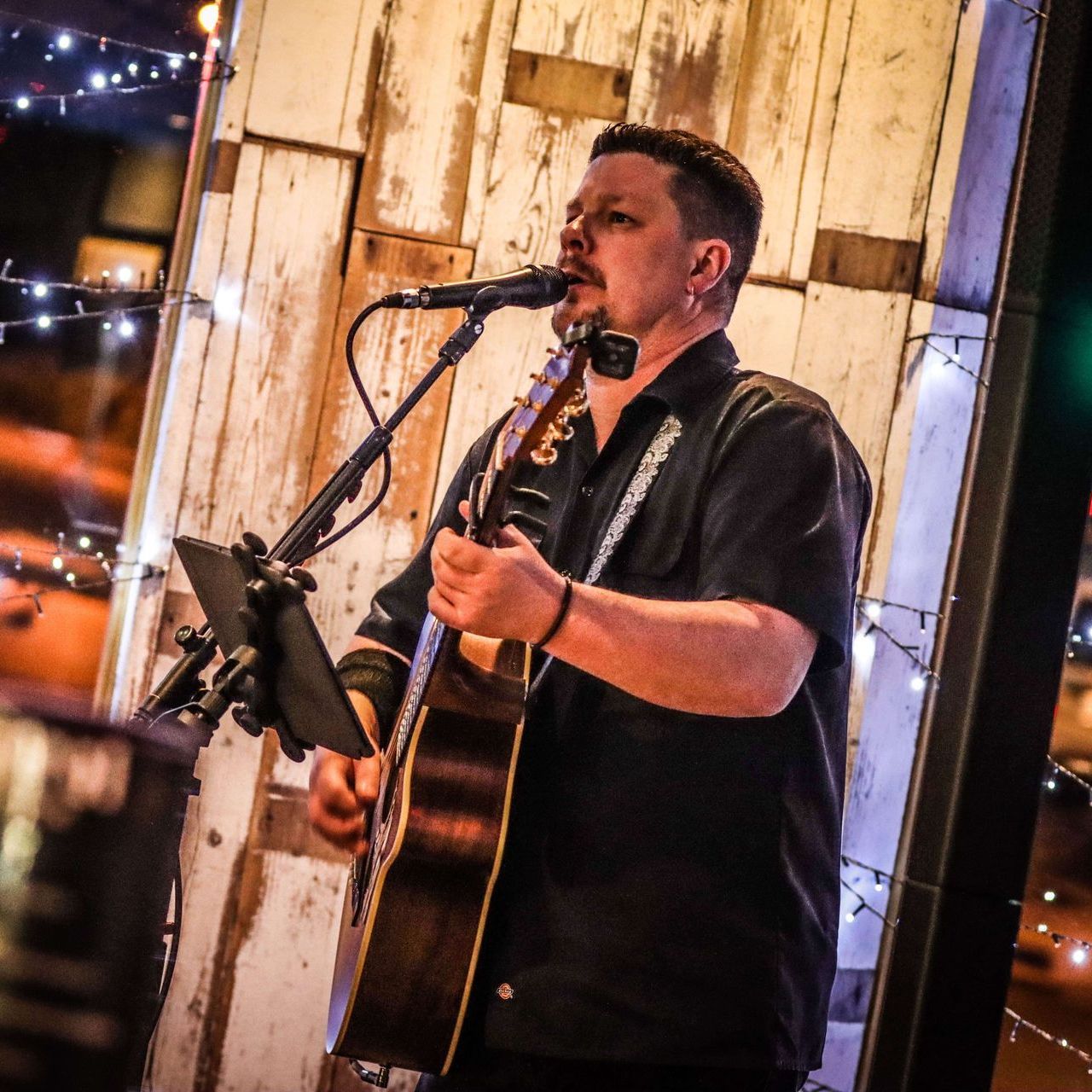 Man playing acoustic guitar and singing into a microphone on a stage. Wooden backdrop with fairy lights.