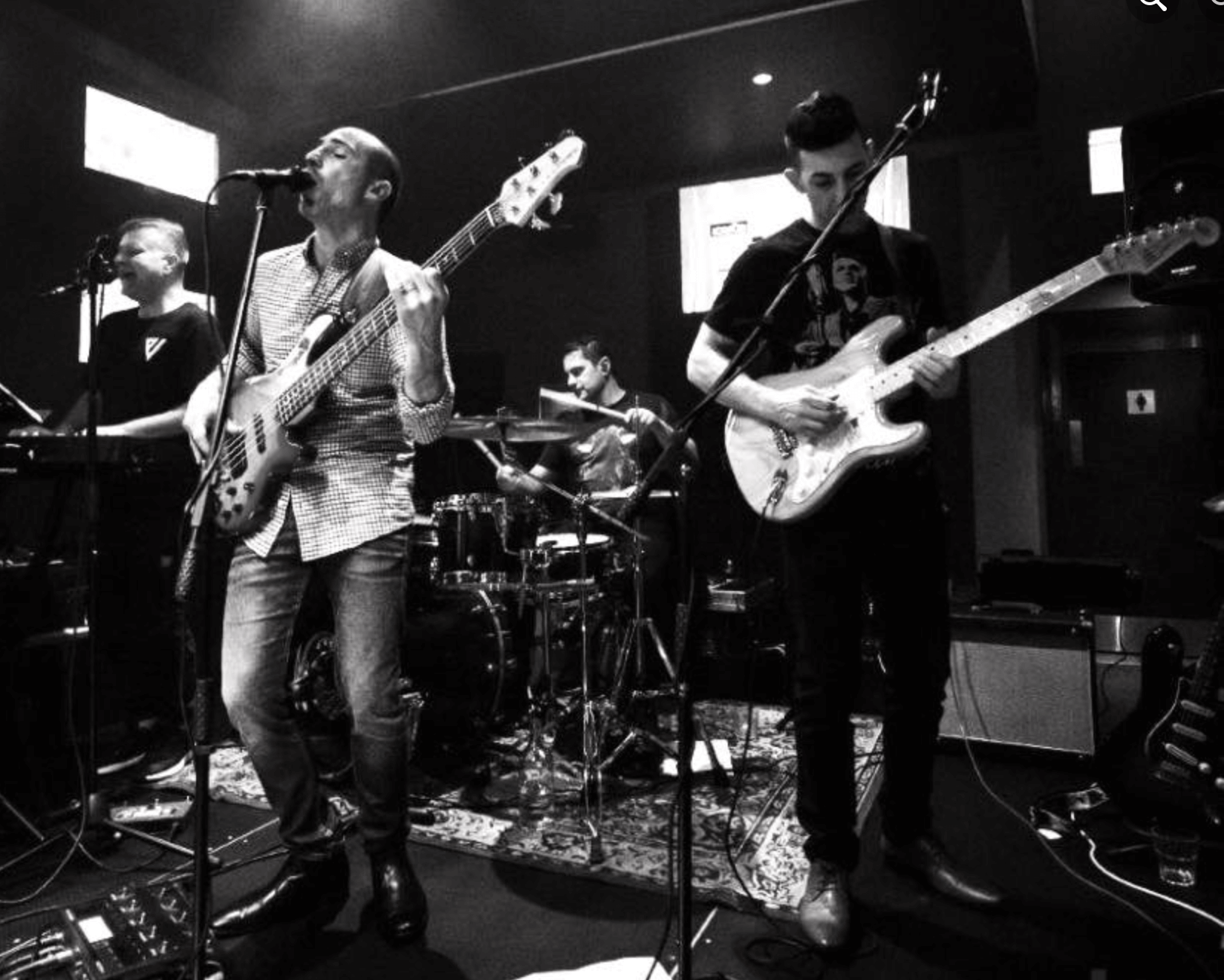 Band performing live on stage: Guitarist, bassist, drummer, keyboardist. Black and white.