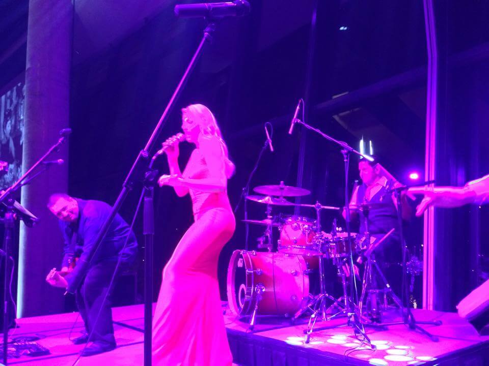 Band performing on stage under purple lights; singer in long dress, drummer, guitarist, and other musicians with microphones.