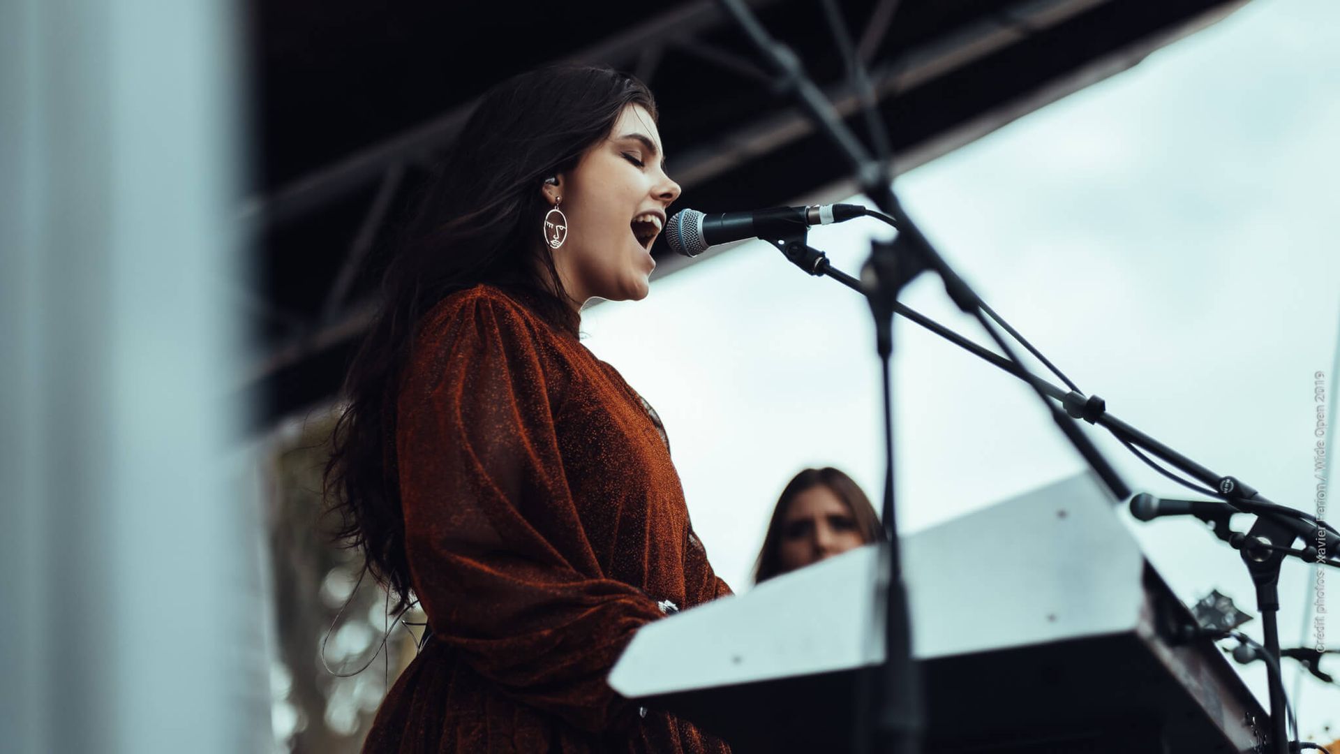 A musician in a brown long-sleeved dress plays a keyboard and sings into a microphone on a stage.