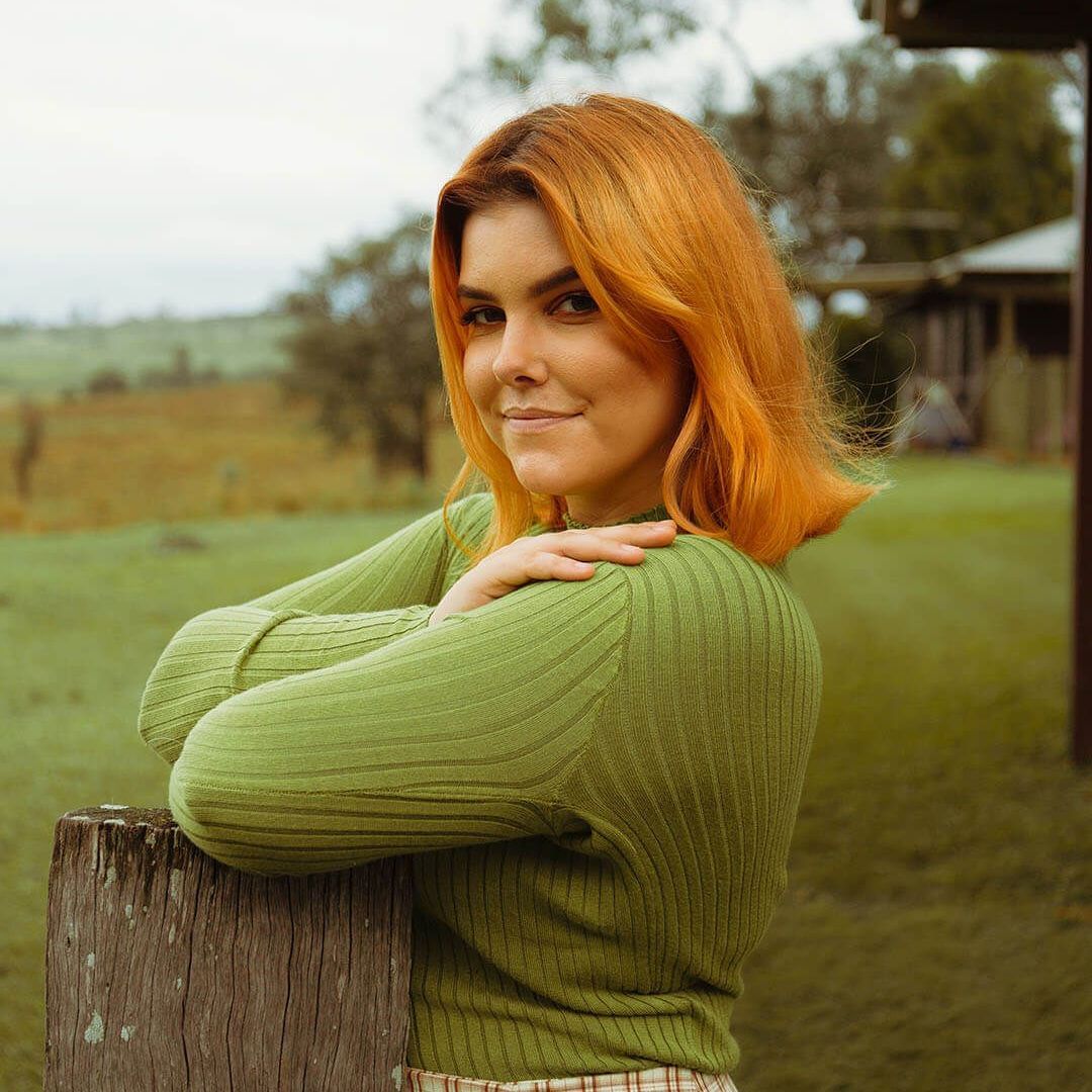 A person with ginger hair in a green sweater leaning against a wooden fence in a grassy field.