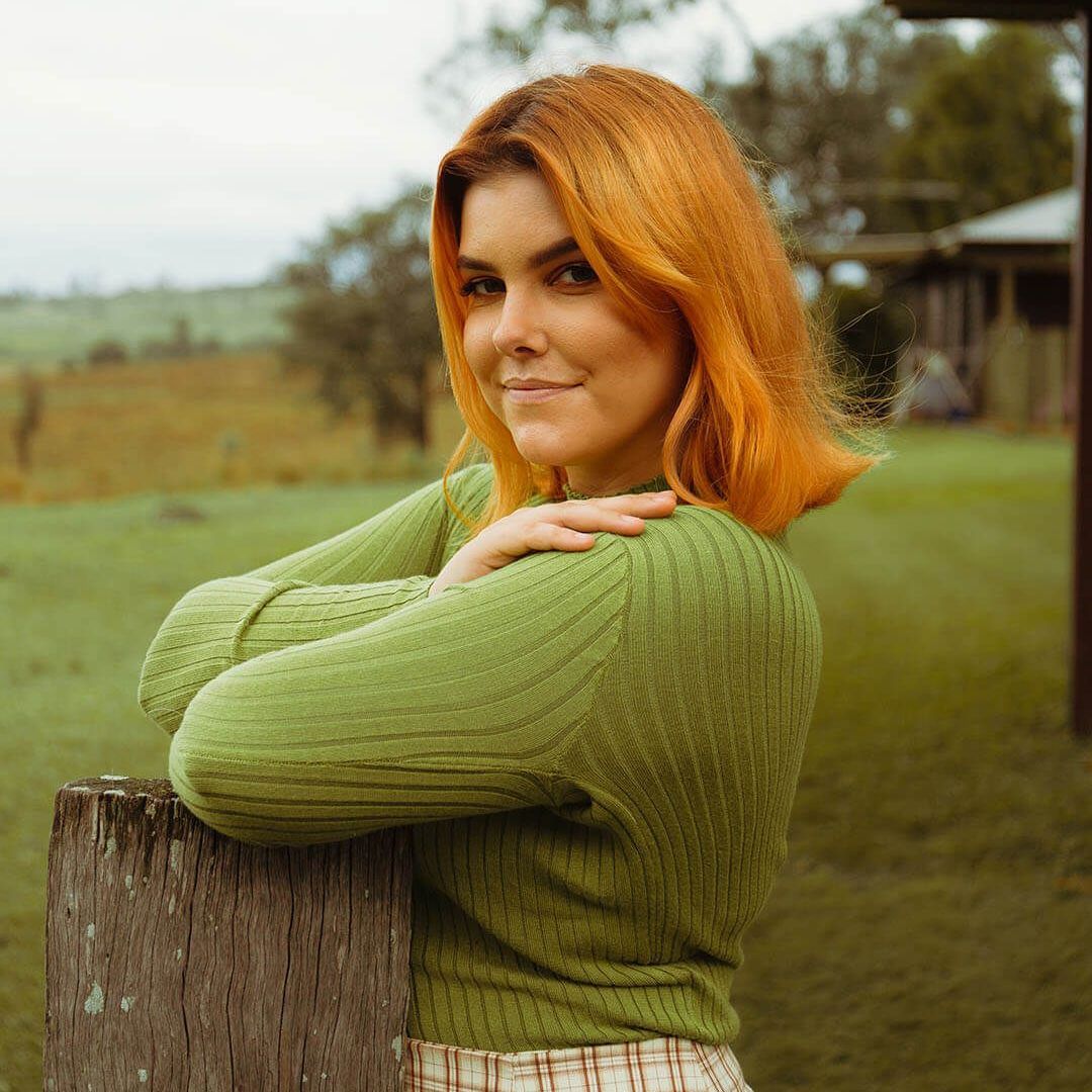 A person with copper-colored hair in a green sweater smiles while leaning on a wooden fence post in a grassy field.