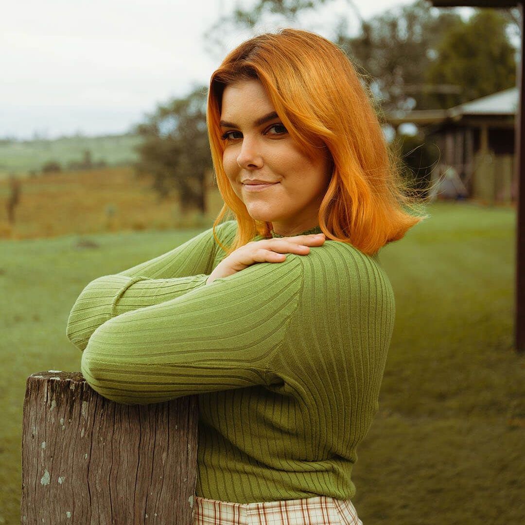 Woman with orange hair smiles, resting arms on a wooden post outdoors in a green sweater.
