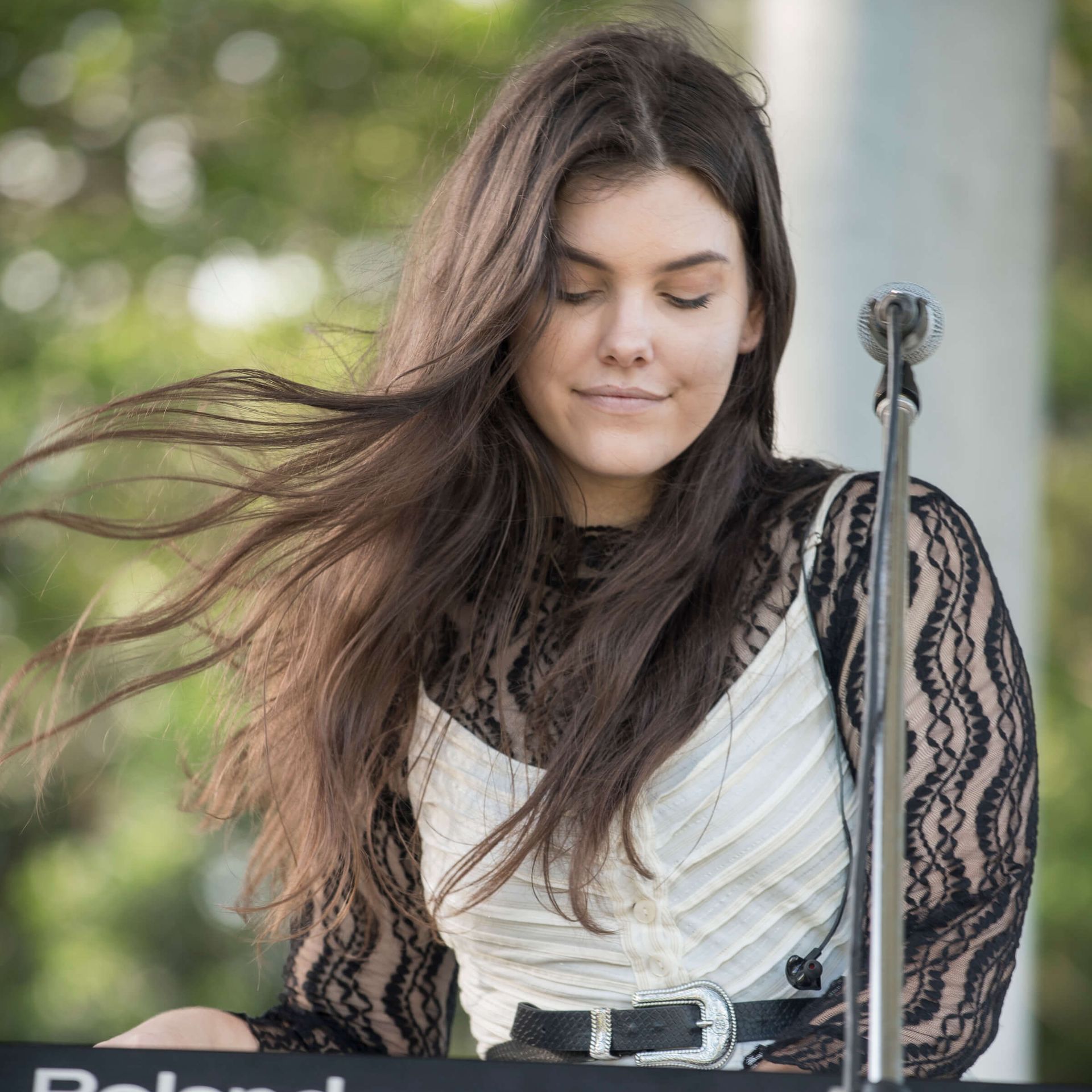 A musician plays a keyboard outdoors, hair blowing in the wind, wearing a black lace top over a white pleated garment.