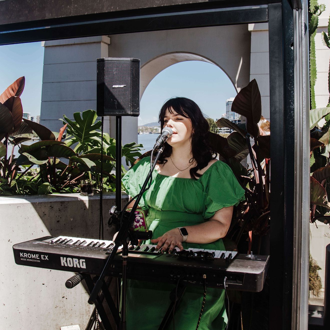 A person in a green dress plays a Korg keyboard and sings into a microphone during an outdoor performance.