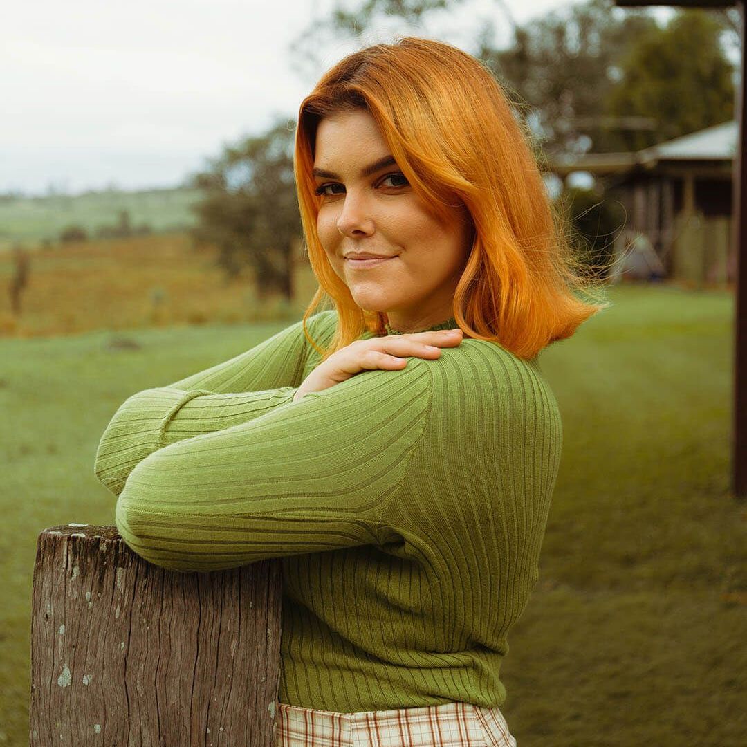A person with ginger hair wearing a green textured sweater, posing outdoors, leaning against a wooden fence post.