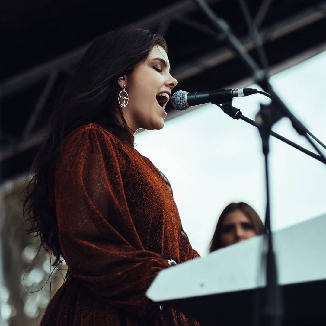 Singer performing at a microphone onstage, wearing a rust-colored top