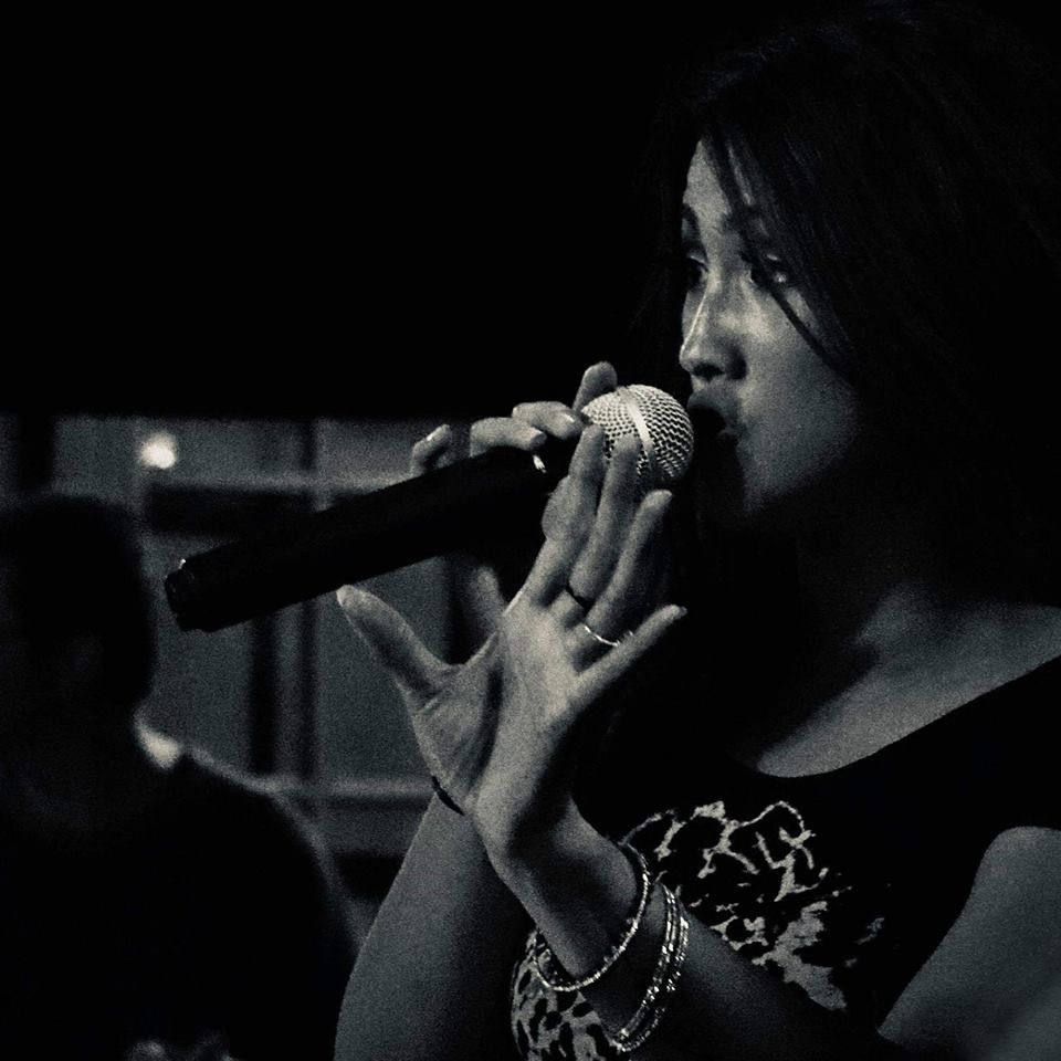 A person in a black patterned top sings into a microphone, hands held near their face in a low-light, monochromatic setting.