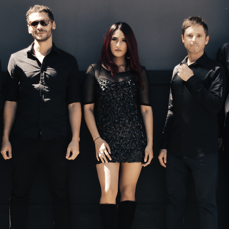 Three people in black attire stand in a row against a solid dark gray wall, posing for a portrait.