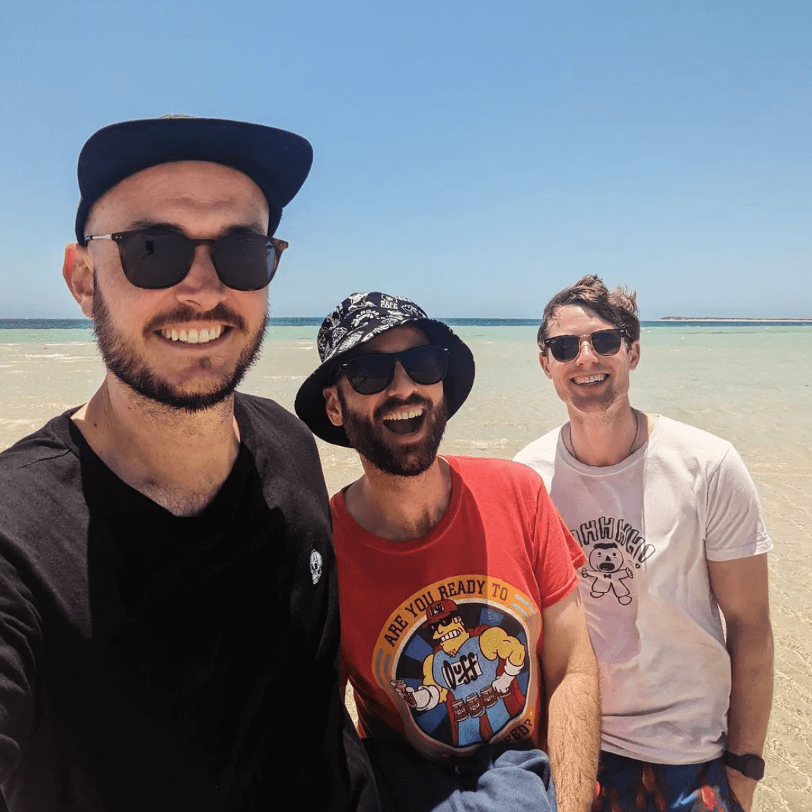 Three people smile while posing for a selfie on a bright, sunny beach by the ocean.