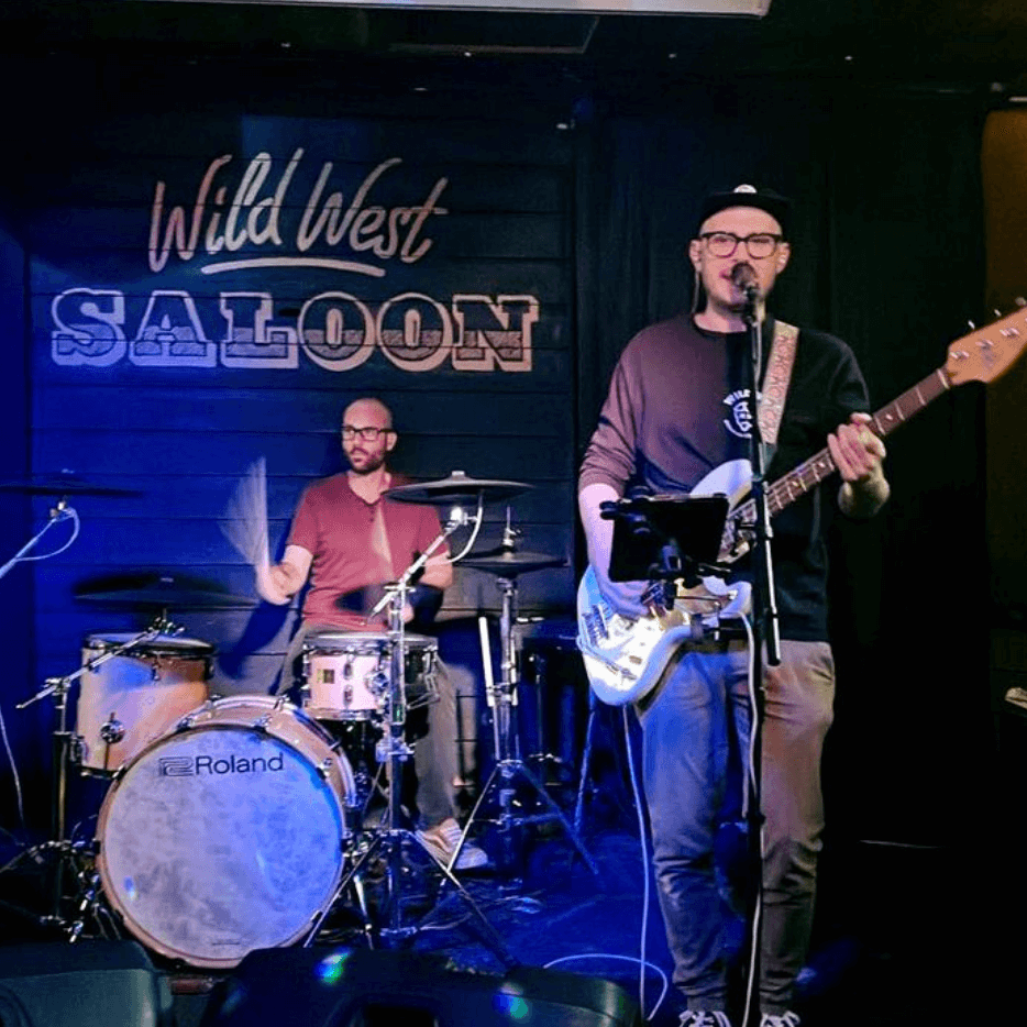 Two musicians perform live on a small stage in front of a 