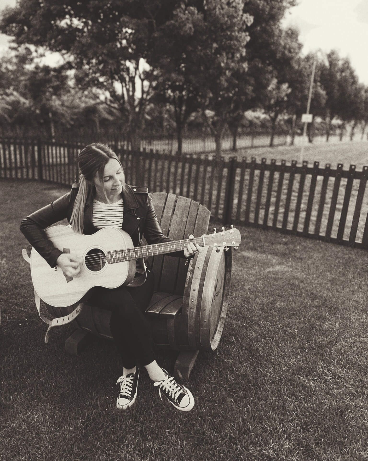 Woman playing guitar while sitting on a barrel chair outside. Wearing a jacket, black pants, and sneakers.