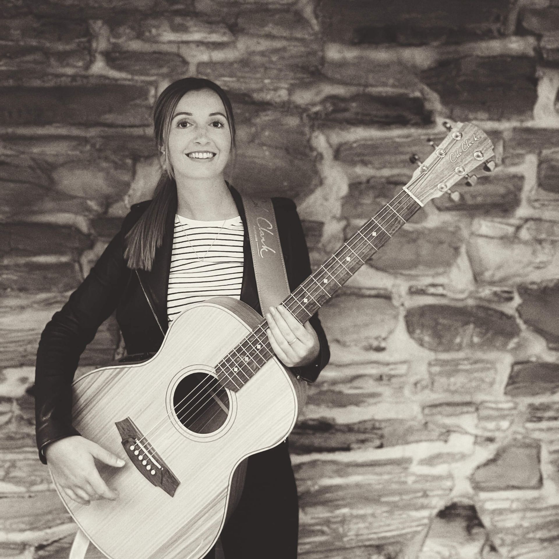Woman holding guitar, smiling, leaning against a stone wall. Black and white photo.