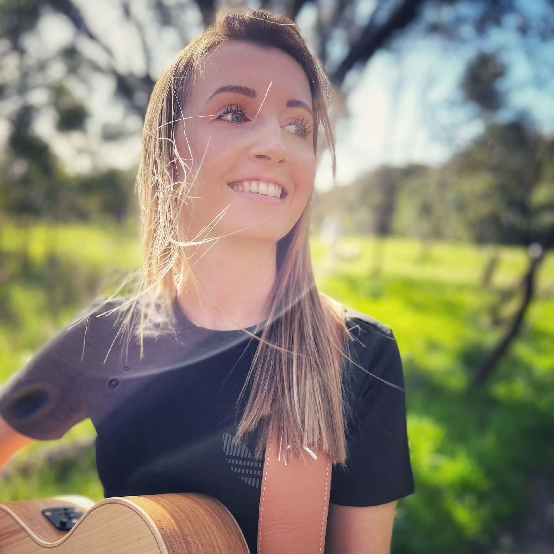 Woman smiling, holding a guitar outdoors, with trees and sunlight in the background.