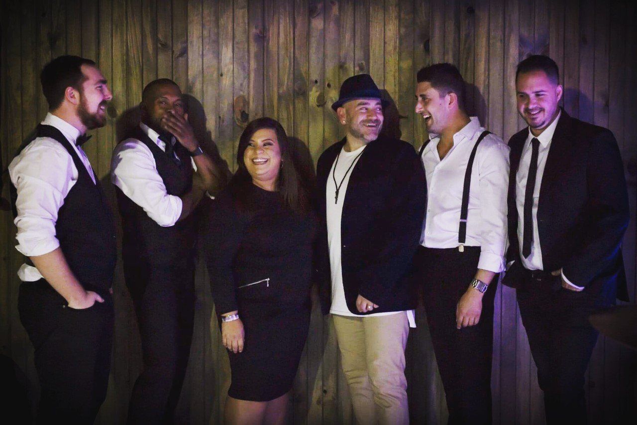 Band of six people smiling, posing in front of a wood-paneled wall.